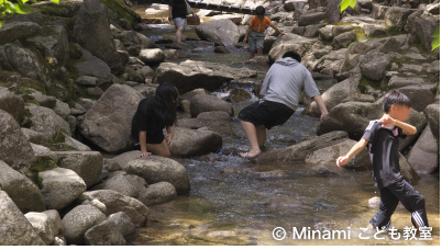 子どもたちが清流にある岩に登ったり、川の水を蹴って遊んだりしている写真